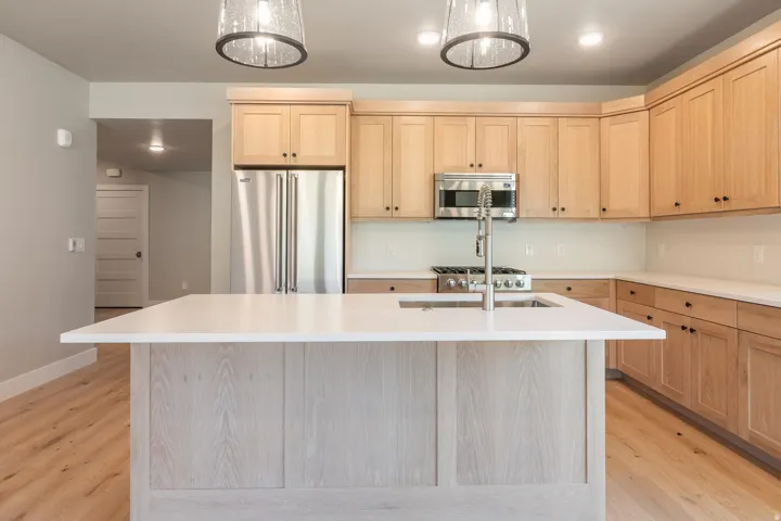Kitchen featuring a central island with white countertop and integrated sink, stainless steel appliances, light wood-finish cabinetry with dark hardware, and wood-finish flooring