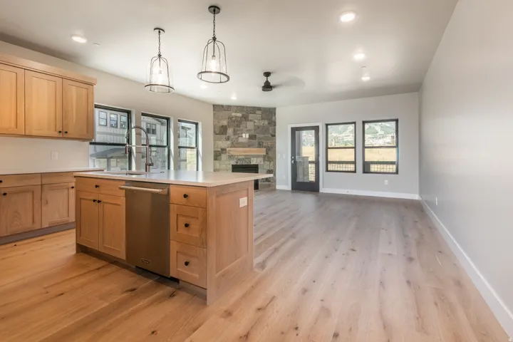 Open-concept living area featuring wood-finish flooring, a kitchen island with integrated sink and dishwasher, light-toned cabinetry, a stone-faced fireplace, and multiple windows with black trim