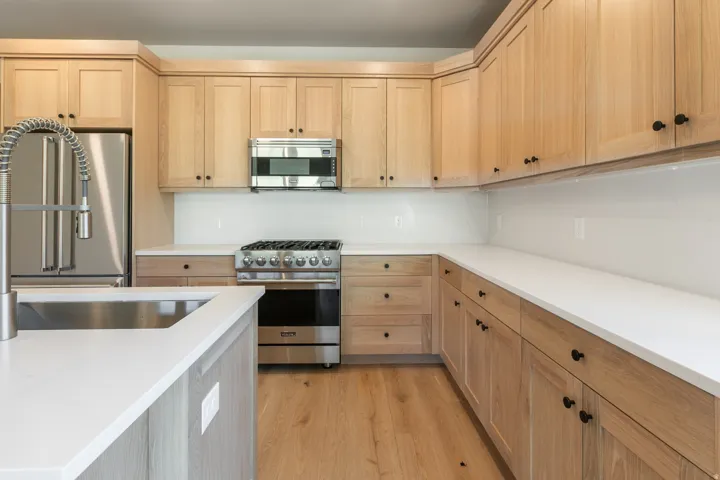 Contemporary kitchen featuring light wood cabinetry, white countertops, stainless steel appliances, a large stainless steel sink with a pull-down faucet, and wood-finish flooring