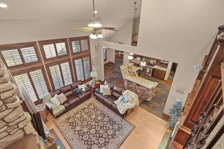 Great room featuring vaulted ceilings, a prominent stone-clad fireplace, large windows with shutters, wood-finish flooring, and an open-concept kitchen with a stone-accented island