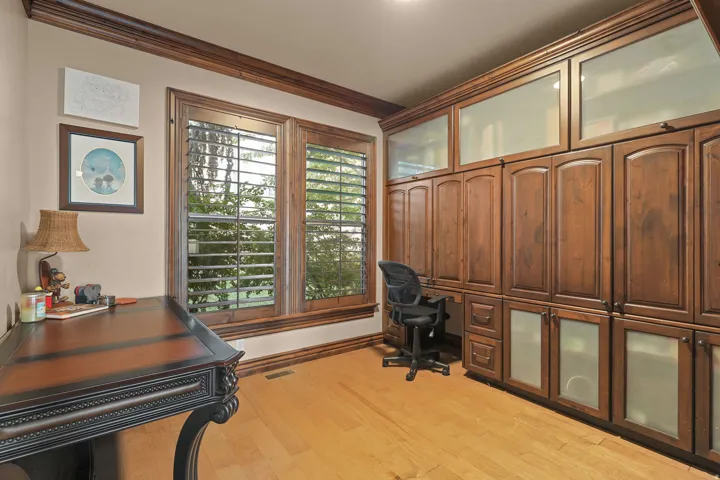 On-suit to bedroom, with built-in cabinetry with frosted glass panels and dedicated desk space, light wood-finish flooring, and multiple windows featuring plantation shutters