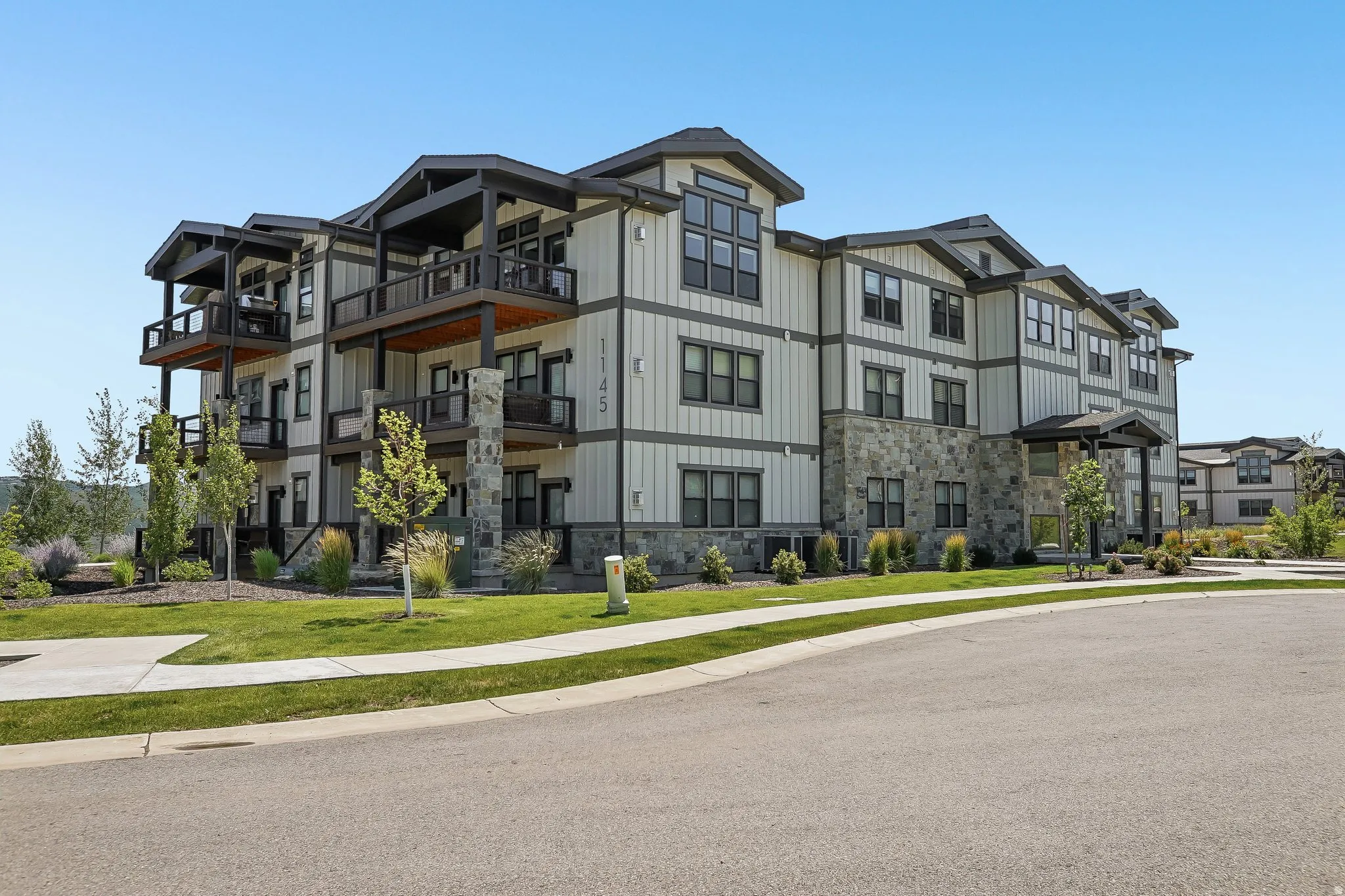 Contemporary multi-story building featuring board and batten siding, stone accents, multiple balconies with glass railings, covered entryways, and manicured landscaping