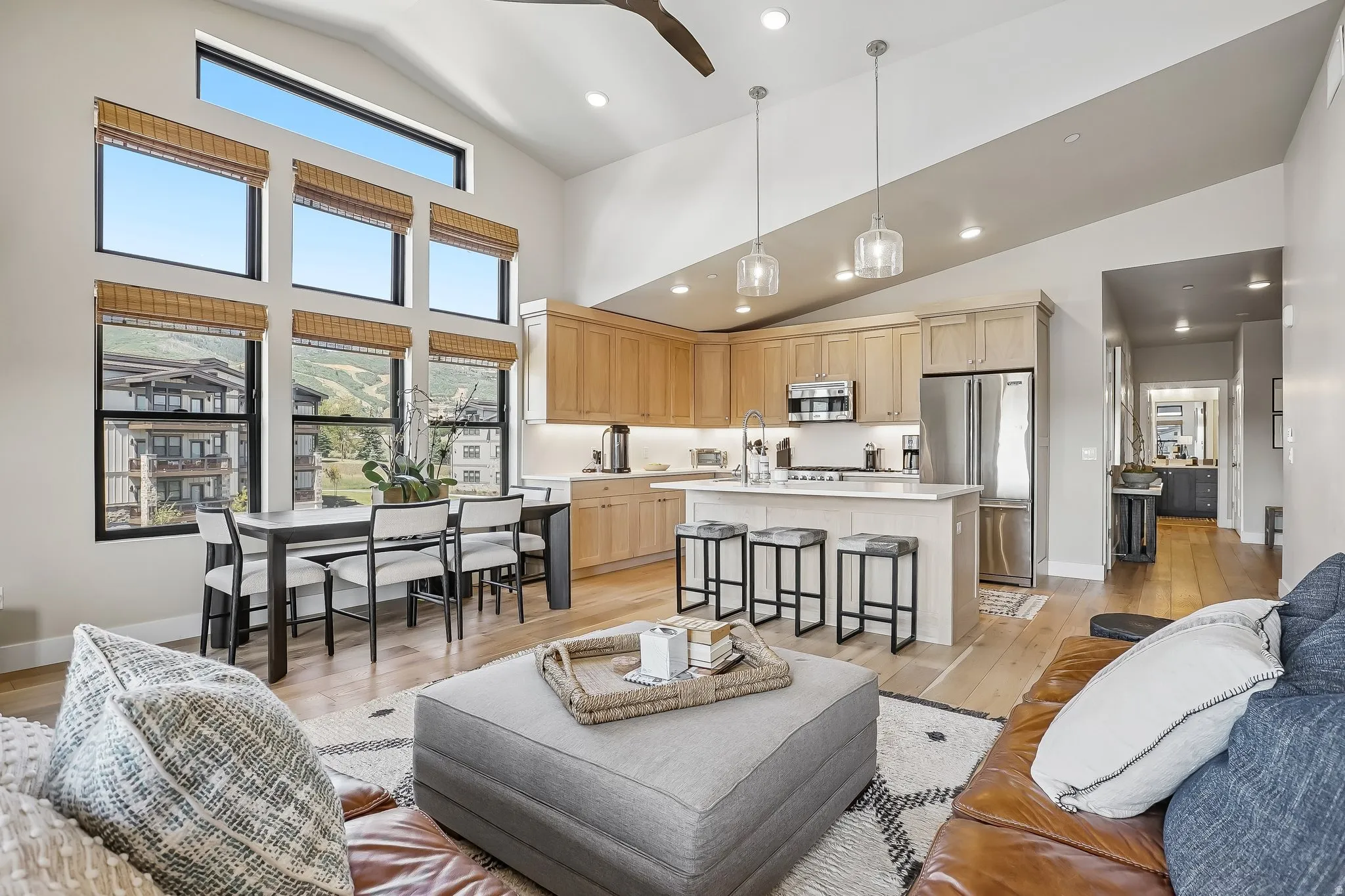 Open-concept living space featuring vaulted ceilings, a full-height window wall, light wood-finish flooring, a kitchen island with seating, and stainless steel appliances