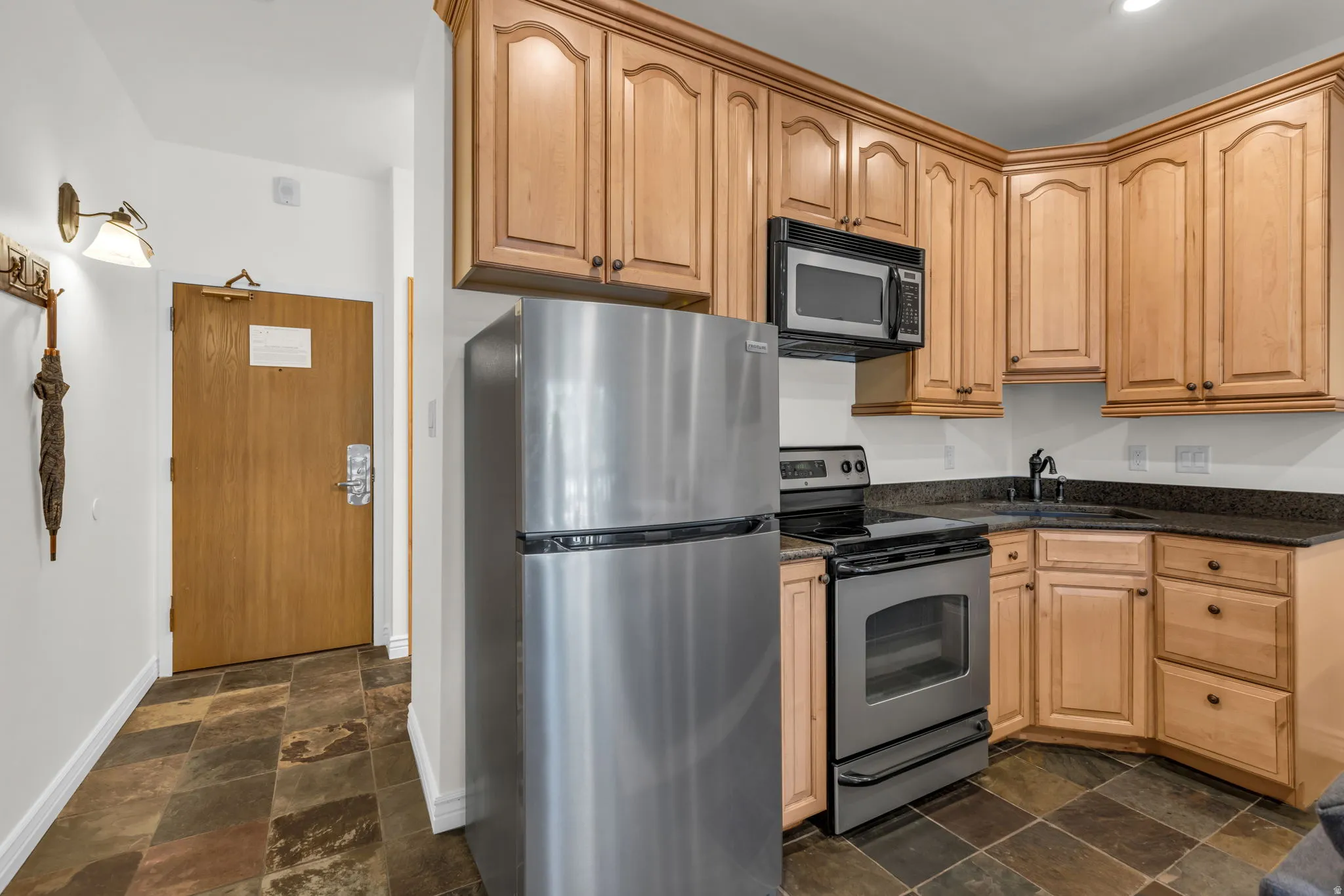 Compact kitchen featuring light wood cabinetry, black countertops, stainless steel appliances, and a corner sink