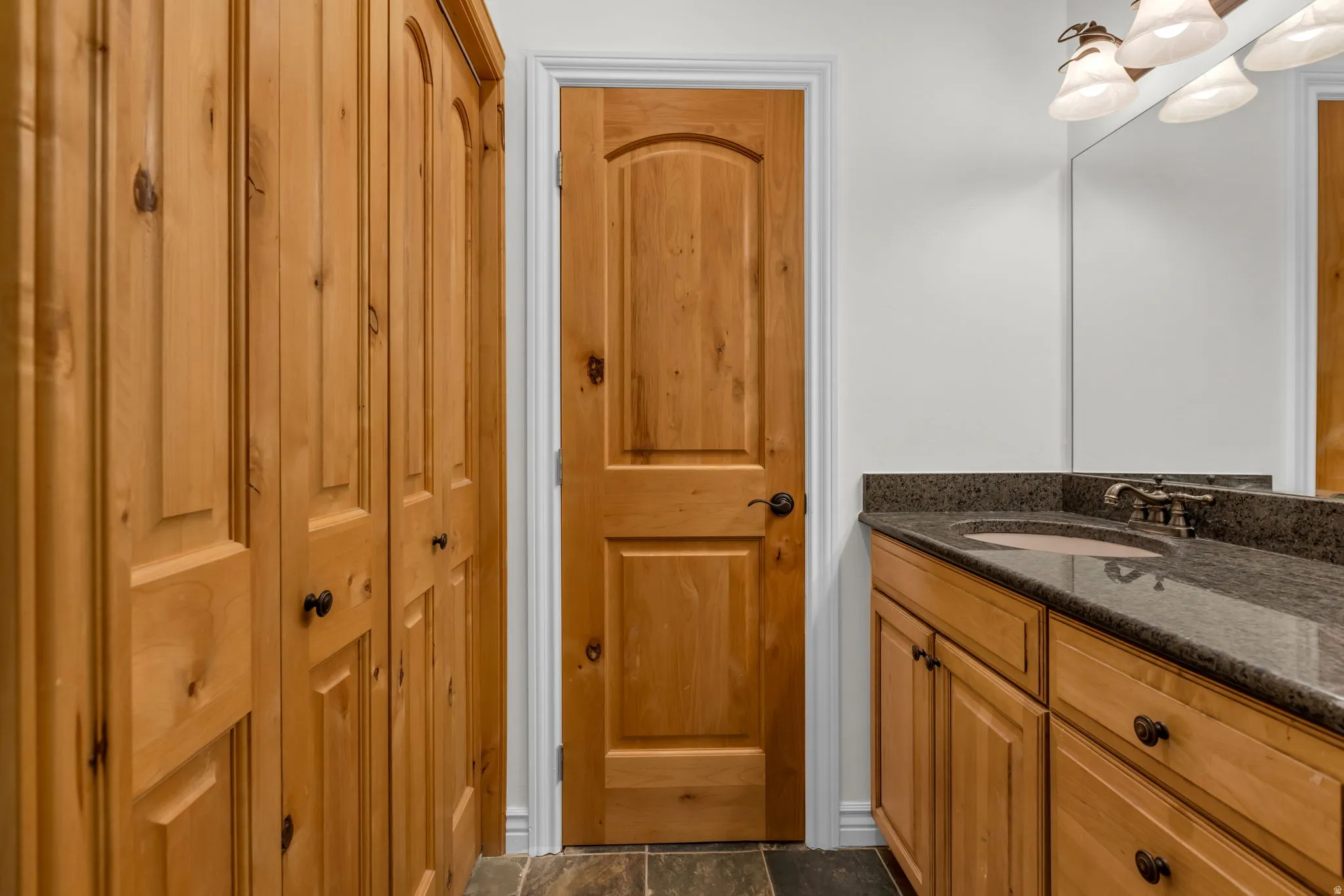 Natural wood cabinetry and door, dark stone countertop with undermount sink, wall-mounted light fixture, and mirrored vanity