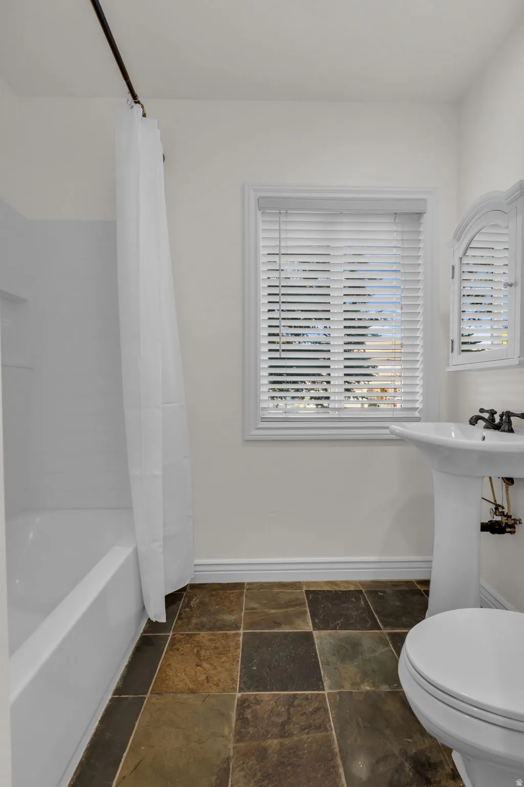 Bathroom featuring a white tub, pedestal sink, toilet, window with blinds, and multicolored square floor tiles