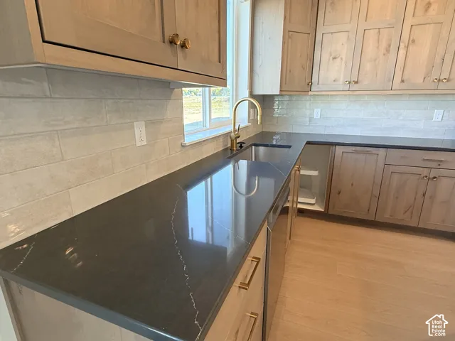 Wood-finish cabinetry with brass hardware, dark stone countertops, undermount sink with brass faucet, horizontal tile backsplash, and wood-finish flooring