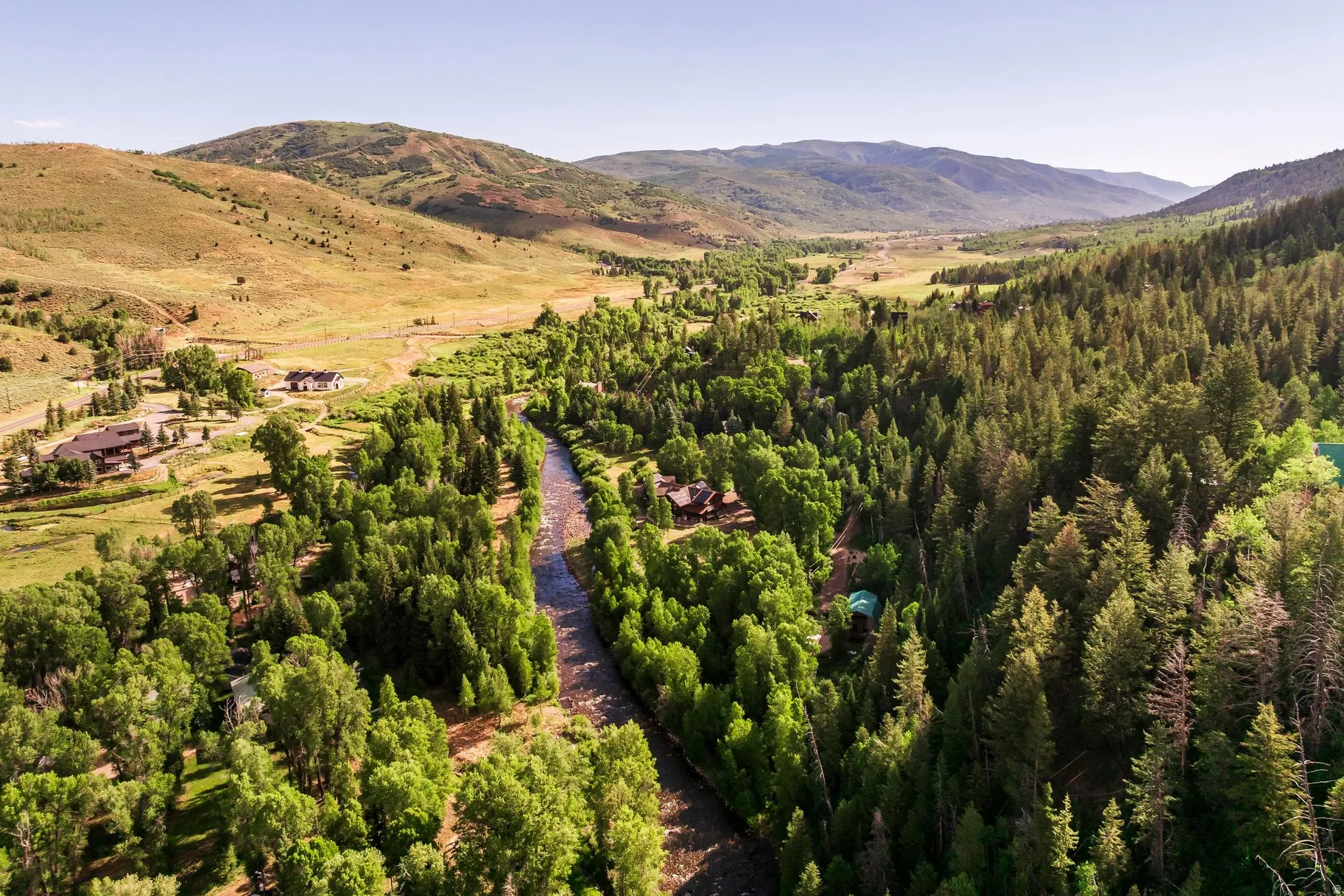 Expansive mountain valley setting featuring a meandering river bordered by dense evergreen and deciduous trees, with scattered properties visible among the foliage and open terrain
