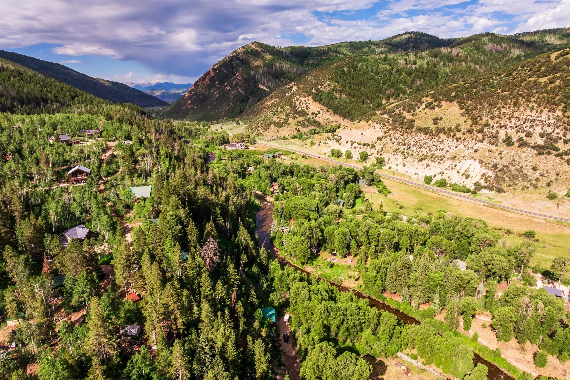 Expansive mountain valley setting featuring a river, dense forest, scattered residences, and a distant road