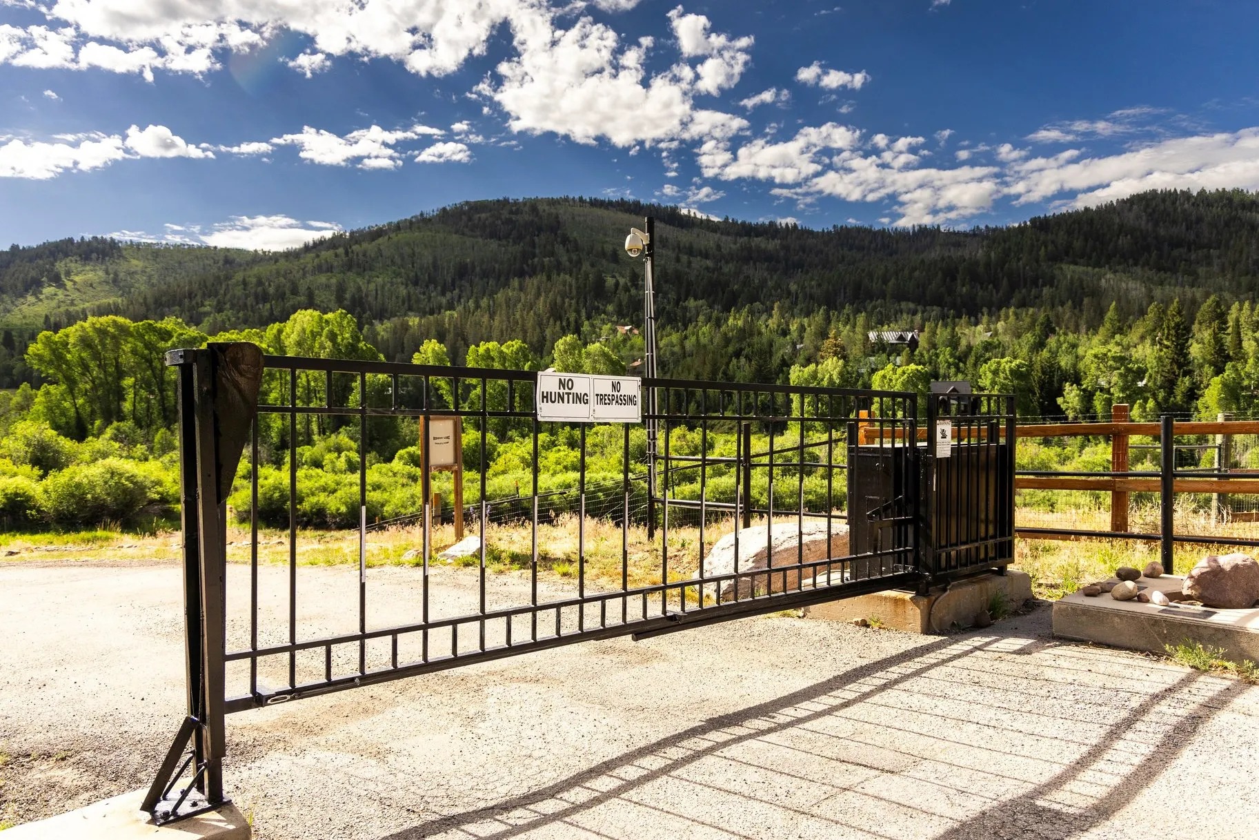 Wrought iron entry gate with security features, set against a backdrop of wooded mountains and mature foliage