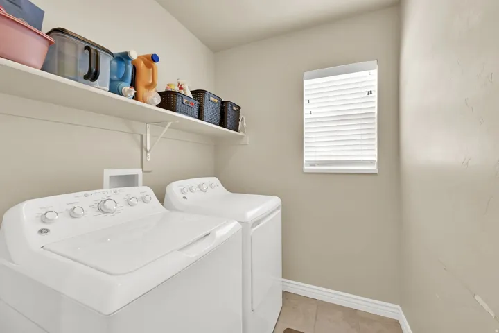 Dedicated laundry space featuring a white top-load washing machine and dryer, an overhead storage shelf, a single window with blinds, and tile flooring