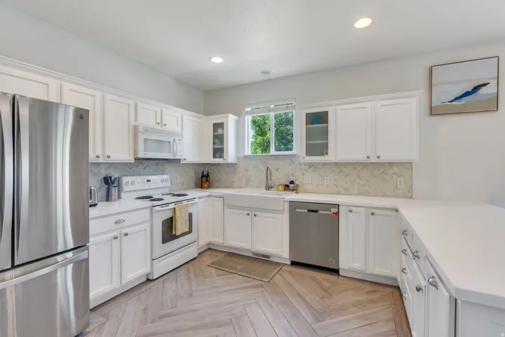 Bright kitchen featuring white cabinetry, light countertops, stainless steel appliances, a white farmhouse sink, and herringbone wood-finish flooring