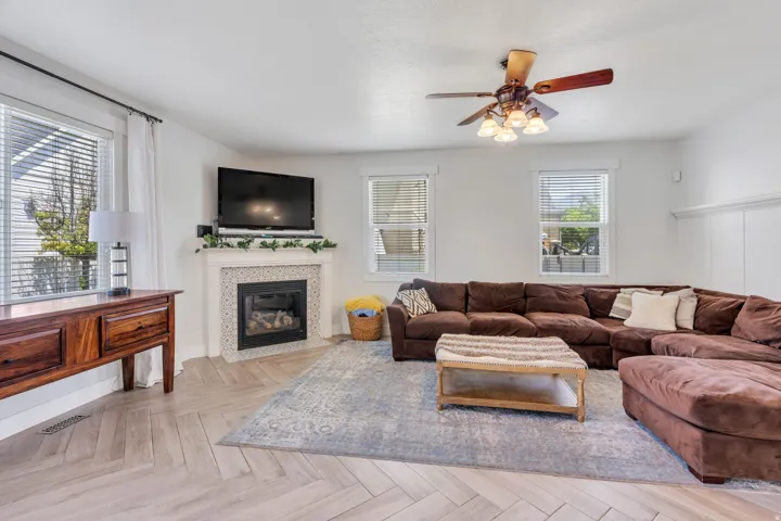 Living area featuring a tiled fireplace with mantel, ceiling fan, multiple windows with blinds, and light wood-finish flooring in a herringbone pattern