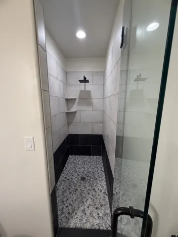 Walk-in shower featuring a glass enclosure with matte black hardware, textured pebble floor, two-tone tile walls with integrated shelving, and a matte black rainfall showerhead