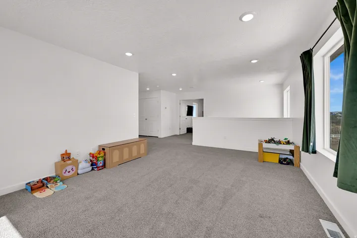 Spacious open loft area featuring plush grey carpeting, recessed lighting, white walls, and a prominent window with a dark green curtain panel