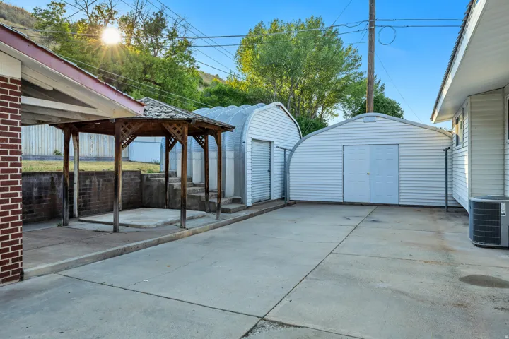 Expansive concrete driveway with a freestanding wooden gazebo, two detached metal storage buildings, and a brick-faced exterior wall