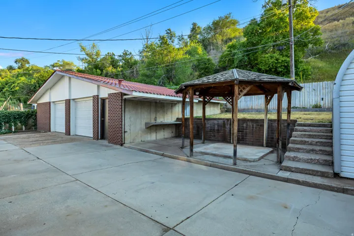 Multi-car garage with white paneled doors and brick accents, concrete driveway, wood-framed gazebo with shingled roof, and concrete steps