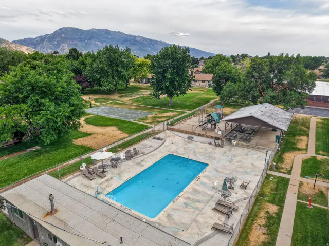 Outdoor swimming pool with surrounding concrete deck, shaded patio with seating, playground area, and a tennis court