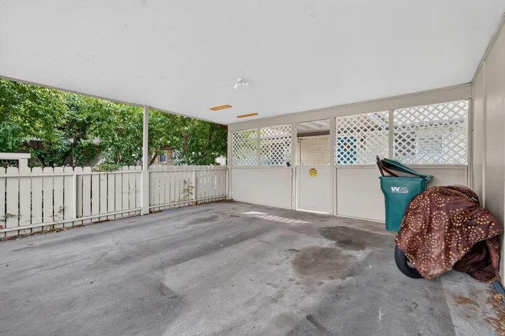 Covered carport with a concrete slab, white lattice paneling, and a white picket fence