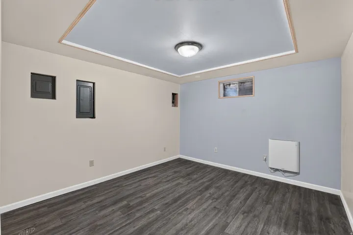Spacious room featuring wood-finish flooring, light blue and beige wall paint, a tray ceiling with a central light fixture, and a wall-mounted heater unit