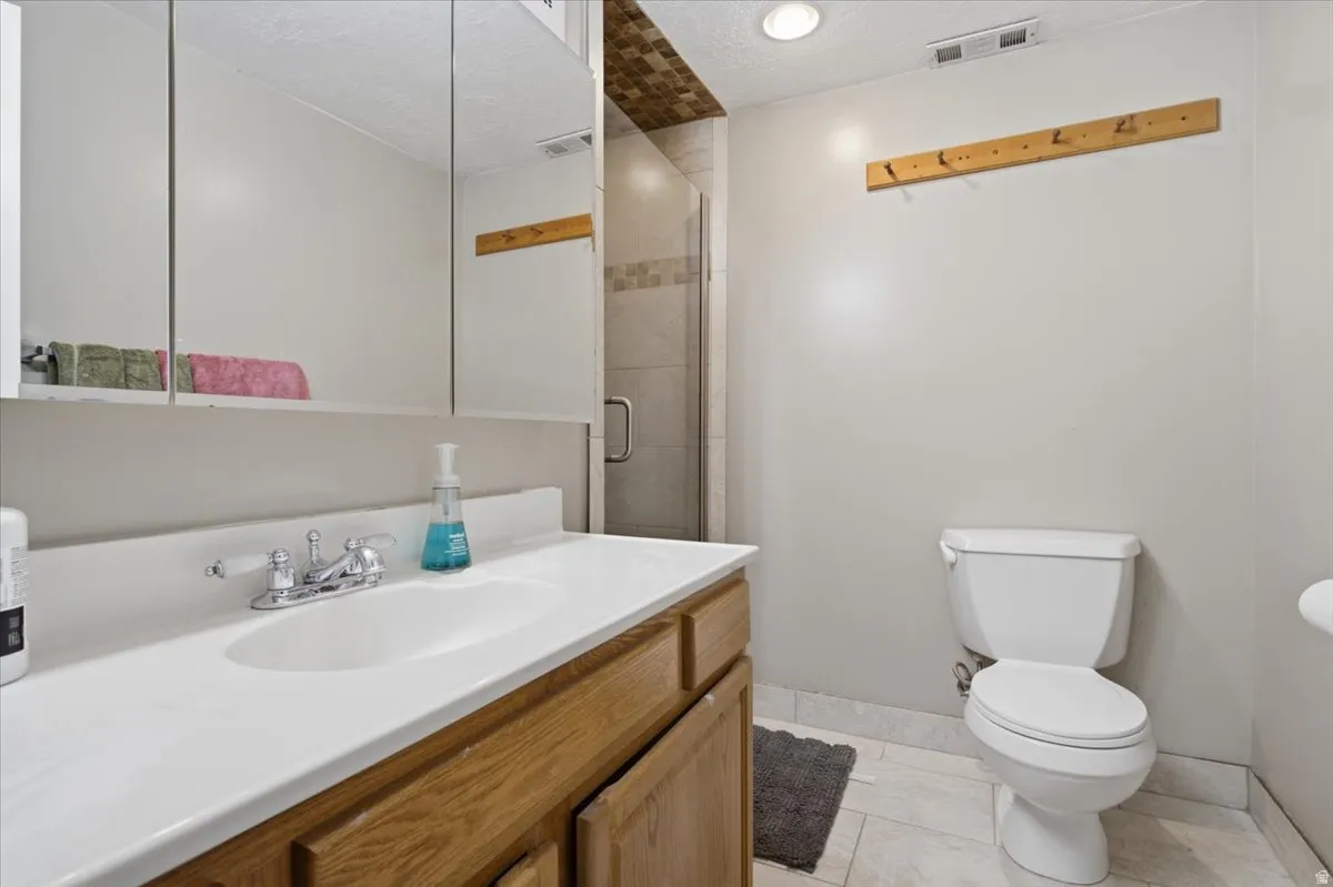 Bathroom featuring a wood-finish vanity with a white countertop and integrated sink, a multi-panel mirrored medicine cabinet, a glass-enclosed shower with tile surround, a toilet, and light-toned tile flooring