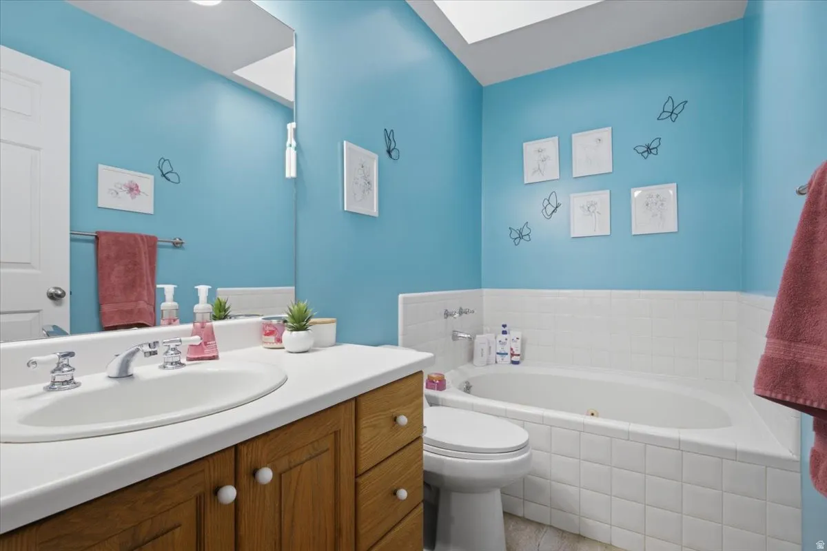 Bathroom featuring a soaking tub with white tile surround, a wood-finish vanity with an integrated sink, a large wall mirror, and a skylight