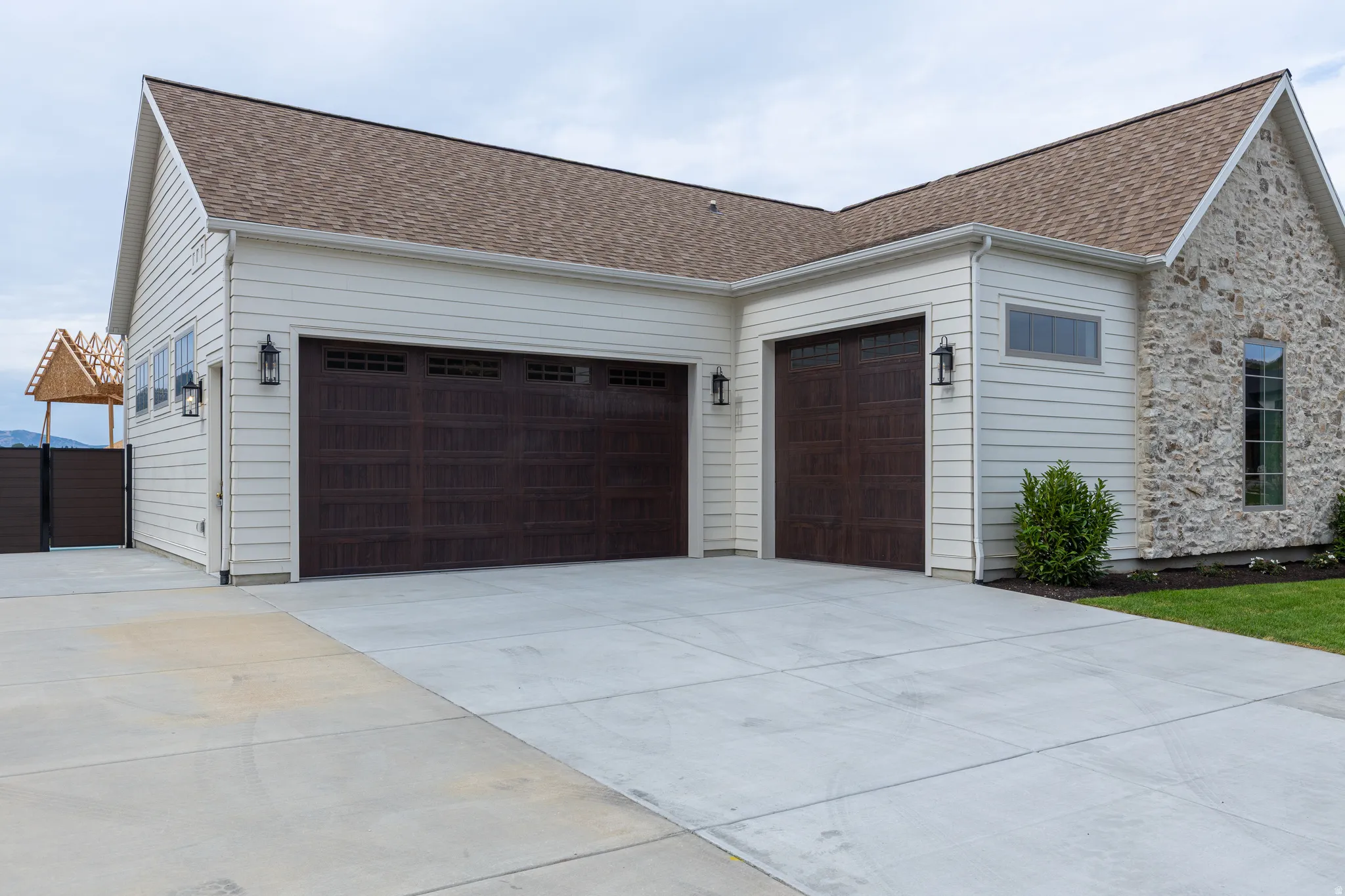 Exterior showcasing a concrete driveway, two dark wood-finish garage doors with decorative windows, white horizontal siding, stone veneer accents, and a brown shingle roof