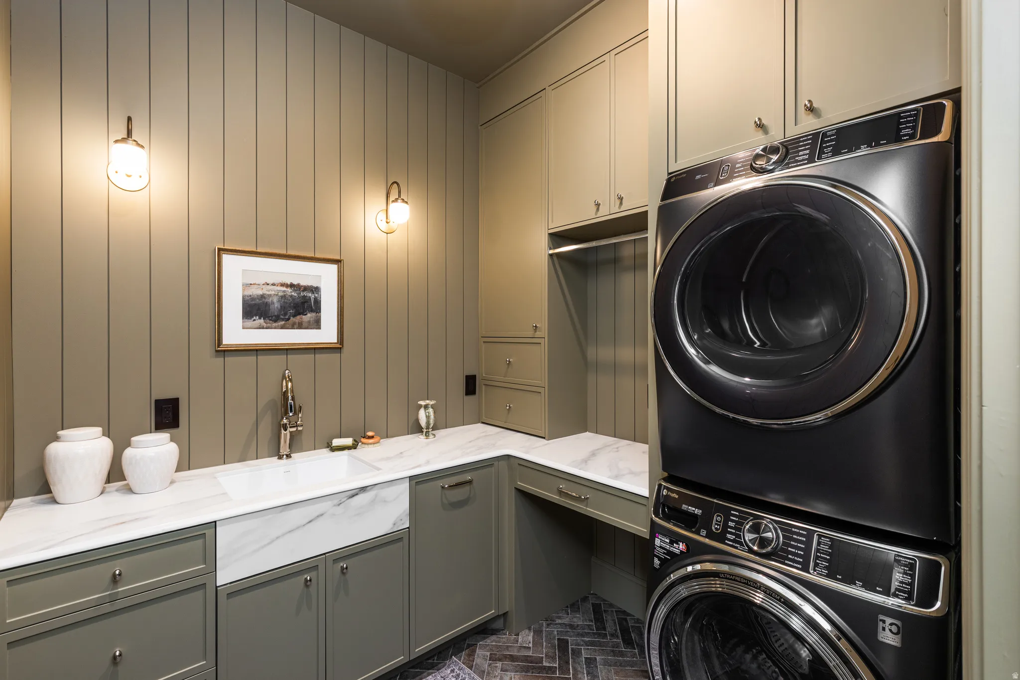 Laundry room featuring vertical paneling, integrated cabinetry, stacked front-load washer and dryer, a farmhouse sink, and a herringbone-patterned floor