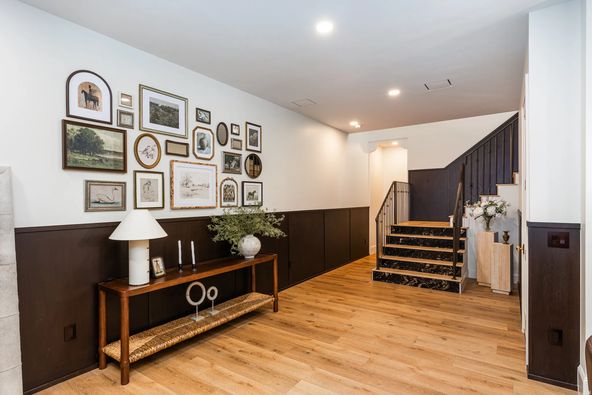 Light wood-finish flooring complements dark wainscoting and a staircase with black risers and wrought iron balusters