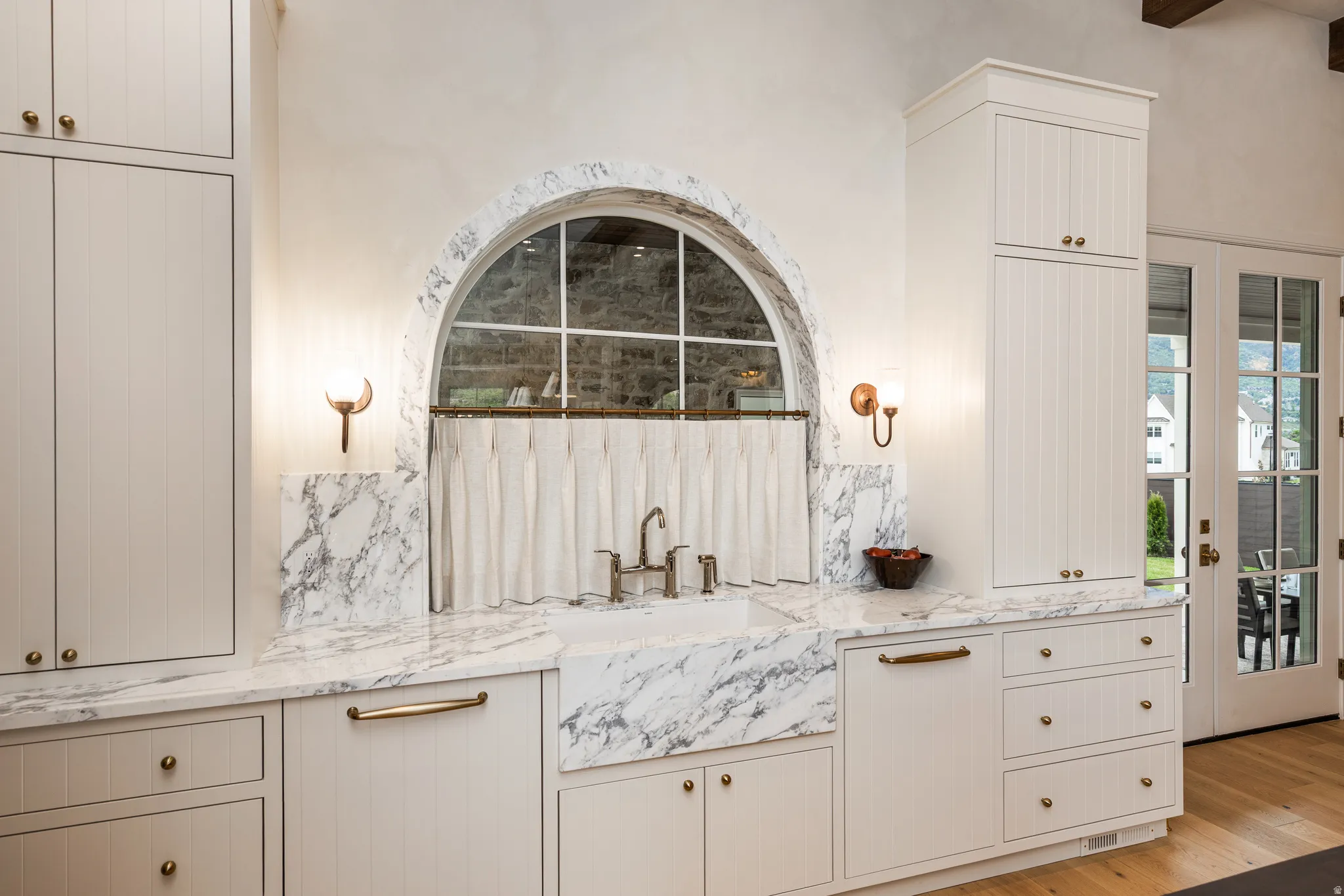 Kitchen workspace featuring an arched window, white cabinetry, marble countertops, an integrated farmhouse sink, and brass hardware