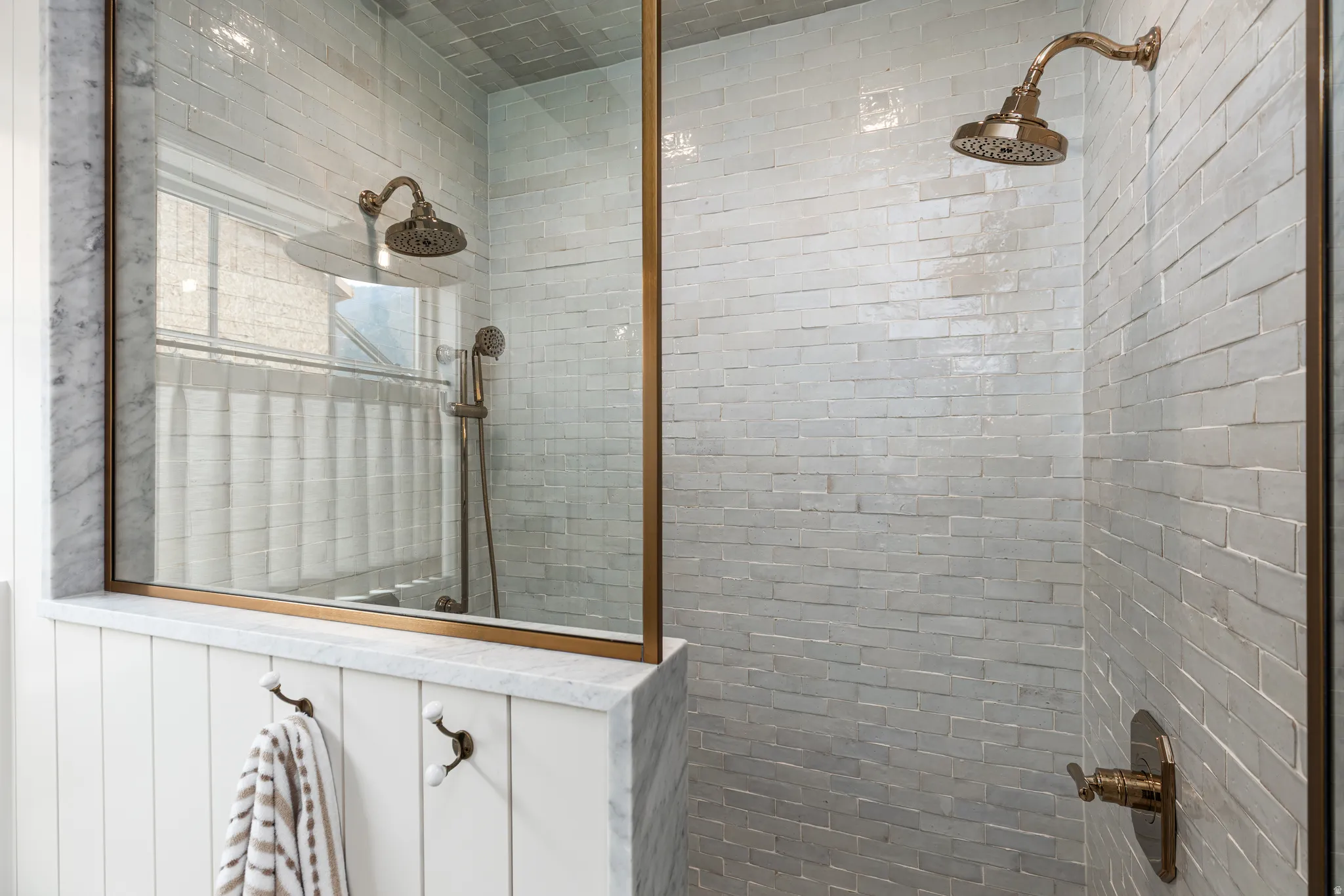 Shower enclosure featuring a frameless glass panel with brass-finish trim, a full-height tiled wall, a rain shower head, a handheld shower fixture, and an integrated marble pony wall