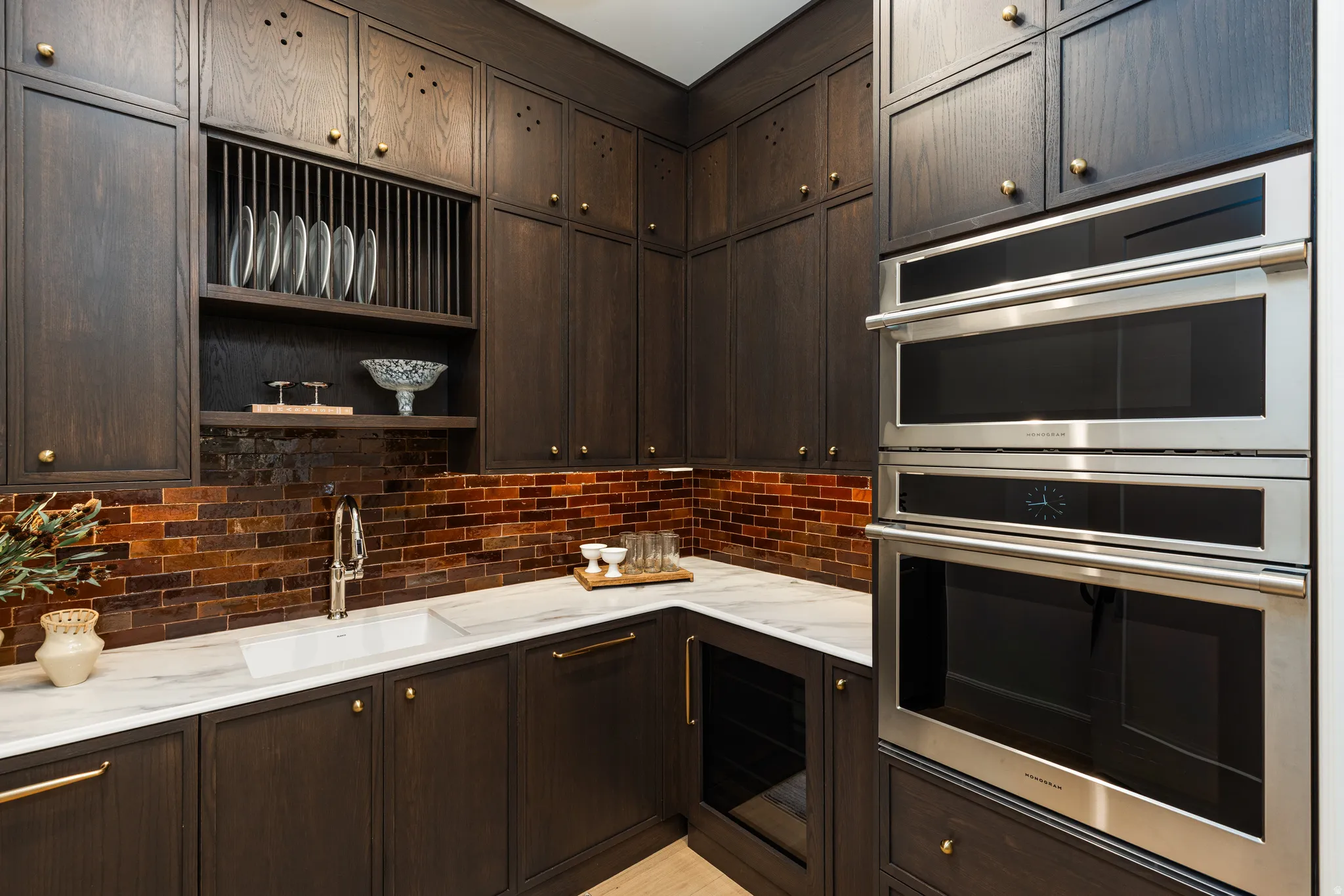 Custom dark wood cabinetry featuring gold-tone hardware, a white undermount sink, marble countertops, an amber glass subway tile backsplash, and integrated stainless steel double ovens