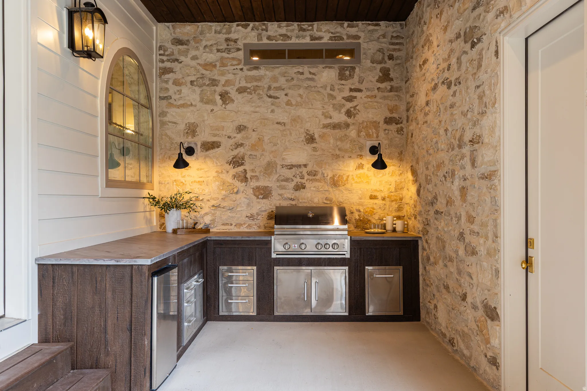 Outdoor kitchen featuring stone wall cladding, built-in stainless steel grill, undercounter beverage refrigerators, and dark wood-finish cabinetry with a stone countertop