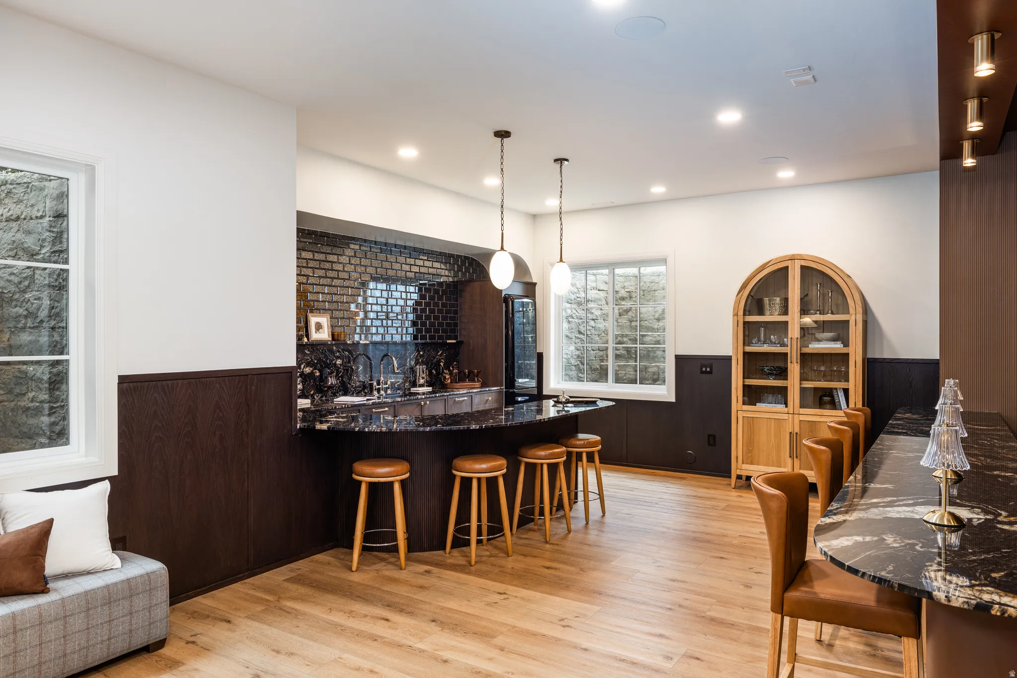 Entertainment space featuring wood-finish flooring, a wet bar with dark countertops and subway tile backsplash, recessed lighting, and two window wells