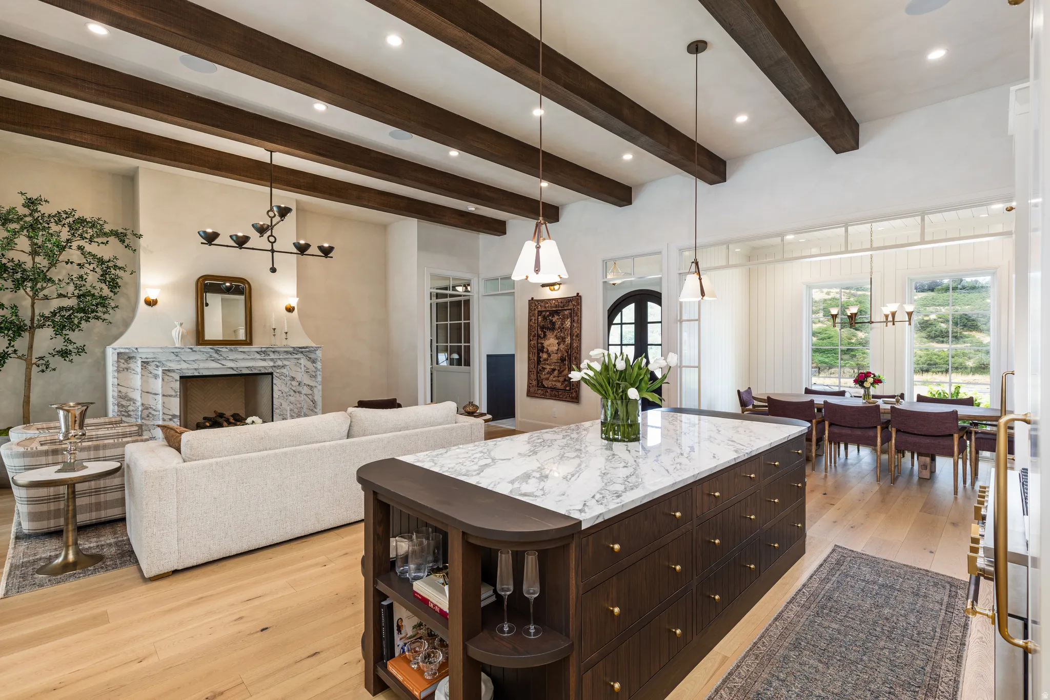 Open-concept living space featuring exposed wood ceiling beams, light wood-finish flooring, and a substantial kitchen island with a stone countertop and integrated storage