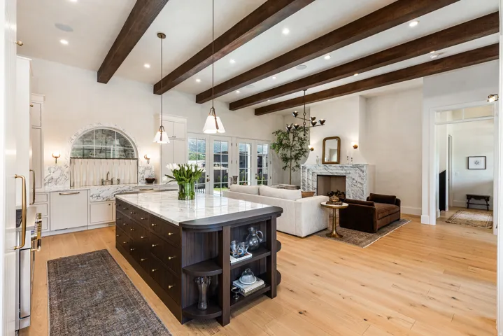 Spacious open-concept living area featuring a large kitchen island with a stone countertop, light wood-finish flooring, exposed ceiling beams, a prominent fireplace with a stone surround, and abundant natural light from multiple French doors