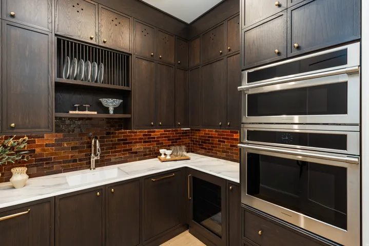Custom dark wood cabinetry featuring gold-tone hardware, a white undermount sink, marble countertops, an amber glass subway tile backsplash, and integrated stainless steel double ovens