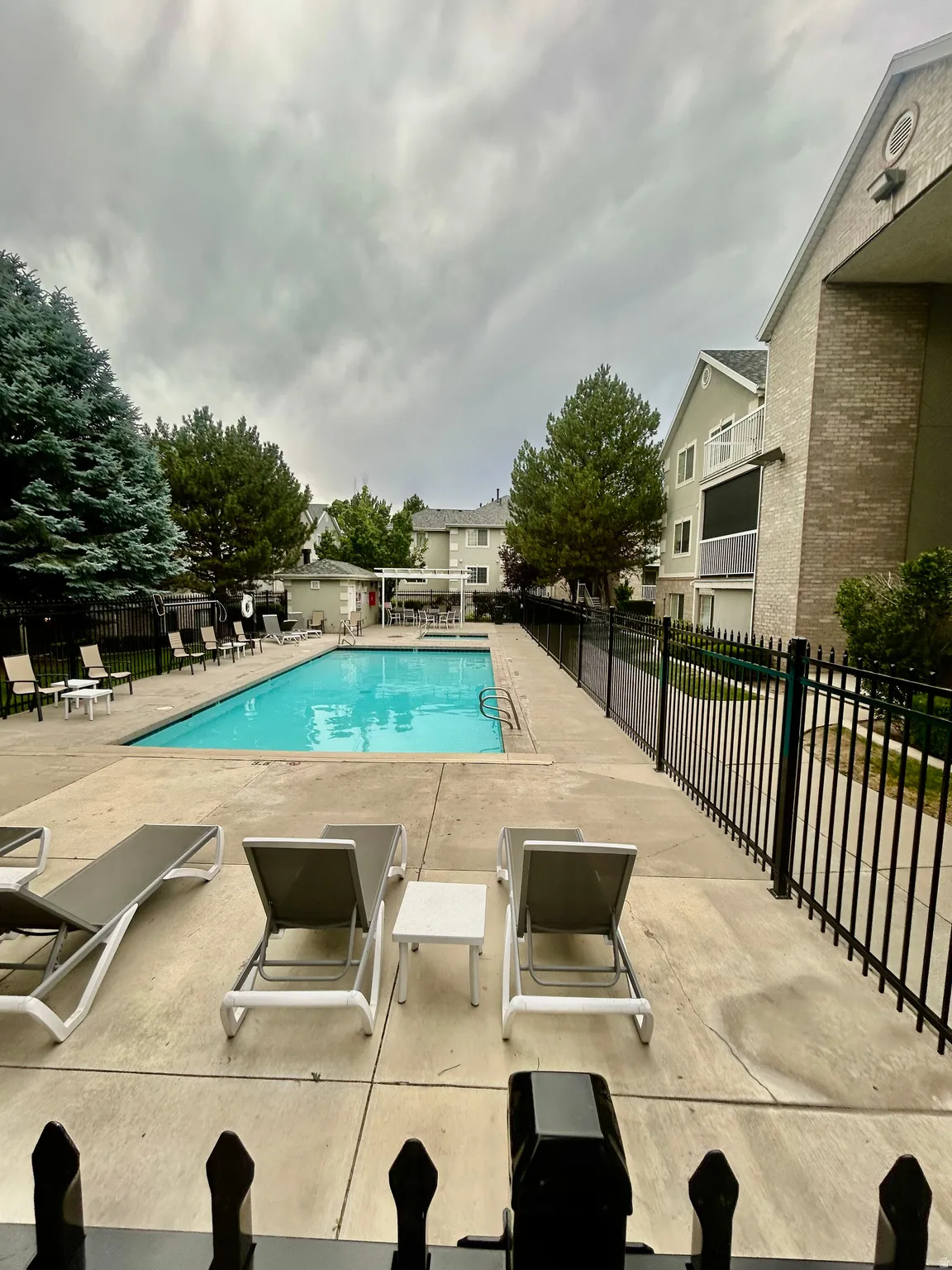 Outdoor swimming pool with surrounding concrete deck, black metal fencing, and mature trees