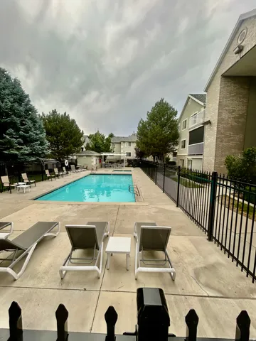 Outdoor swimming pool with surrounding concrete deck, black metal fencing, and mature trees