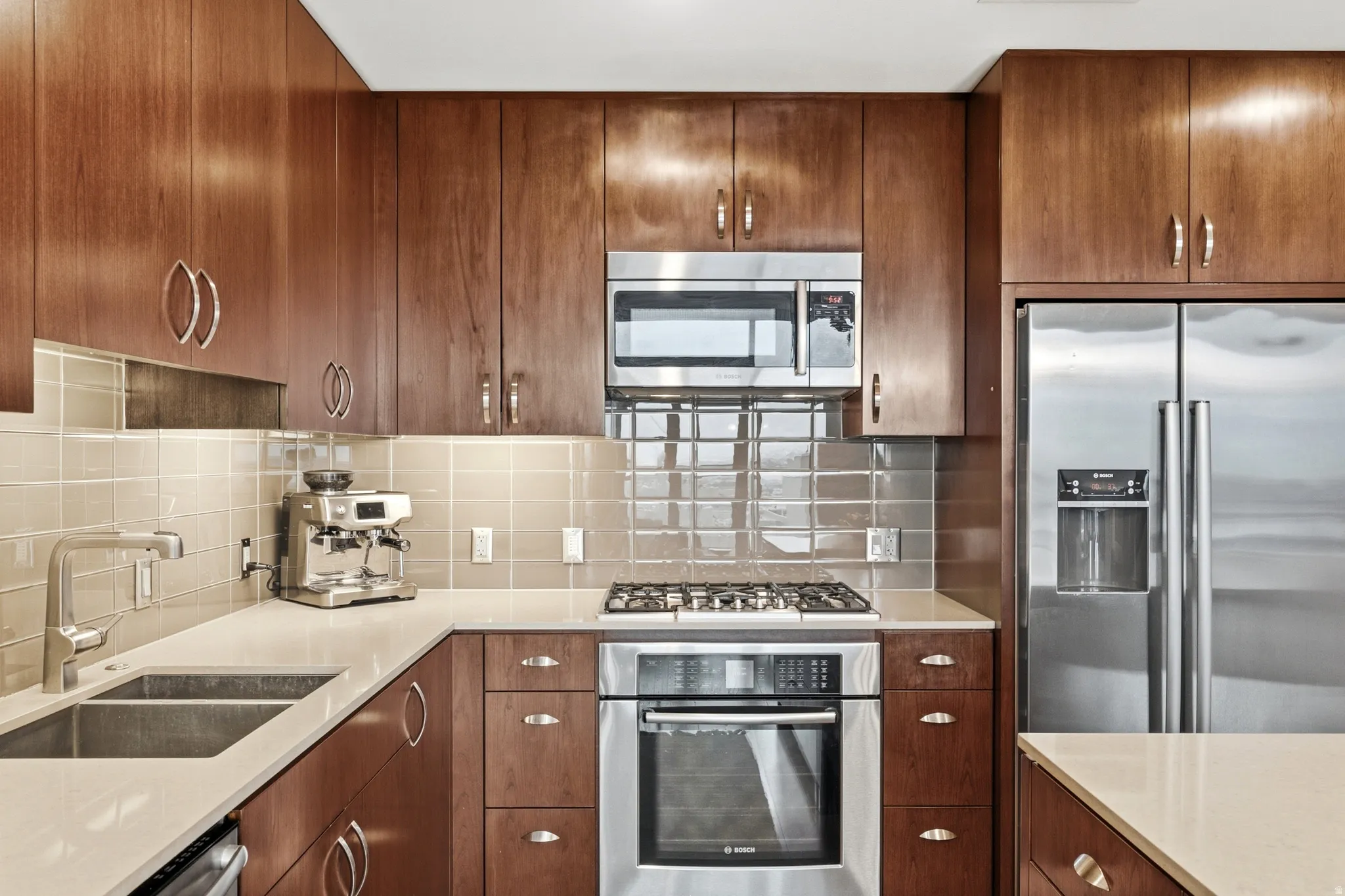 Contemporary kitchen featuring rich wood cabinetry, stainless steel appliances, a double basin sink, light-toned countertops, and a tiled backsplash