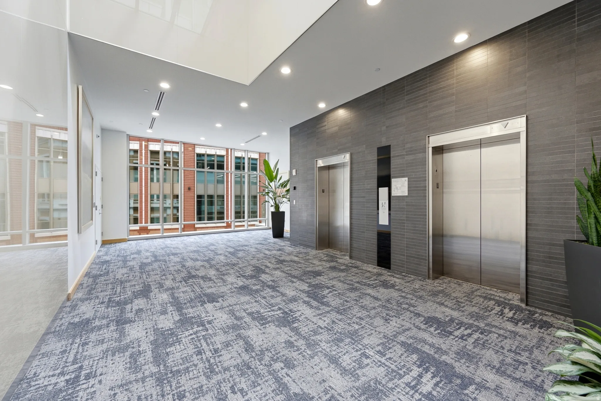 Spacious lobby featuring textured carpet flooring, full-height windows, a gray tiled wall, recessed lighting, and two stainless steel elevators