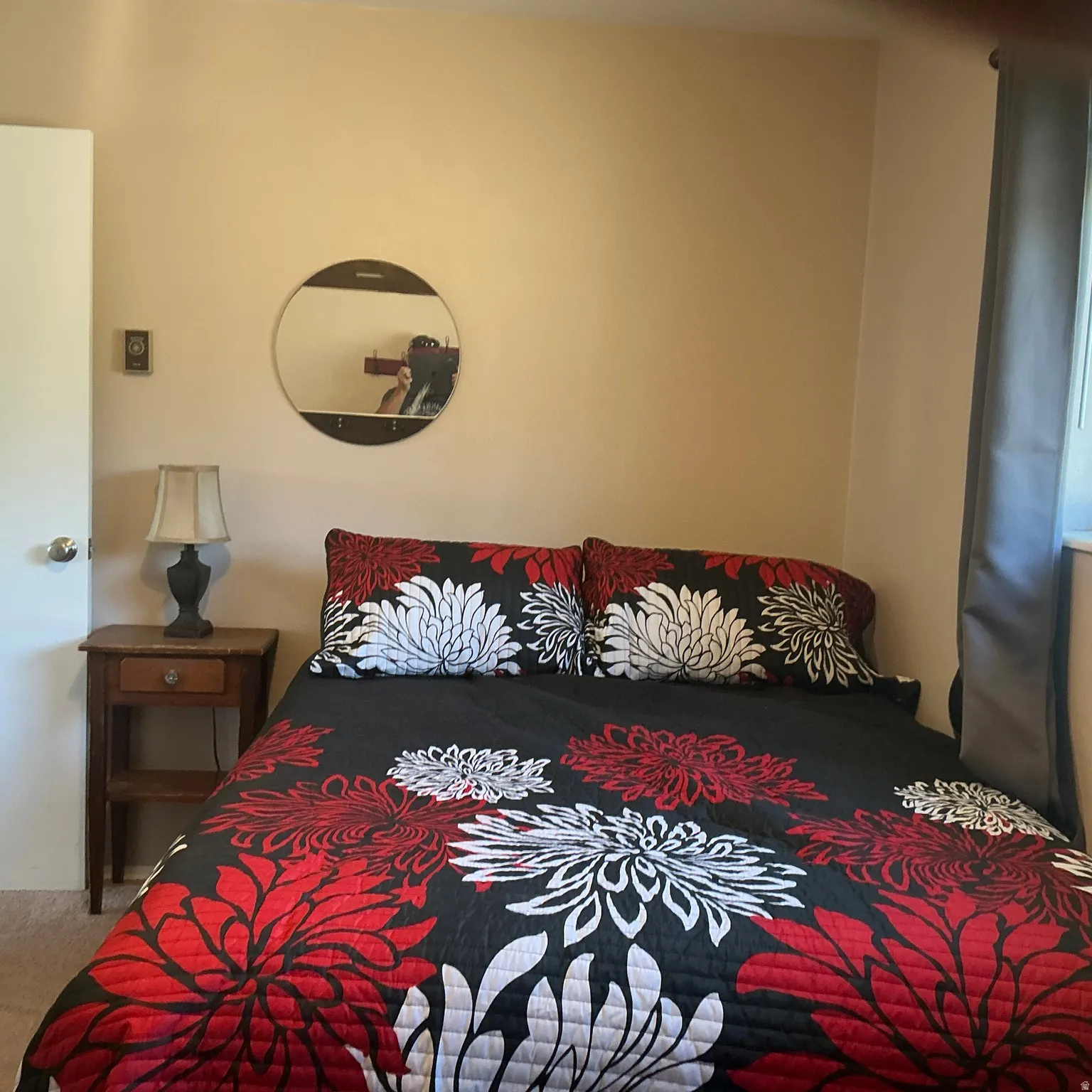 Bedroom featuring neutral wall paint, carpet flooring, a dark wood-finish nightstand, a wall-mounted circular mirror, and a window with dark curtains