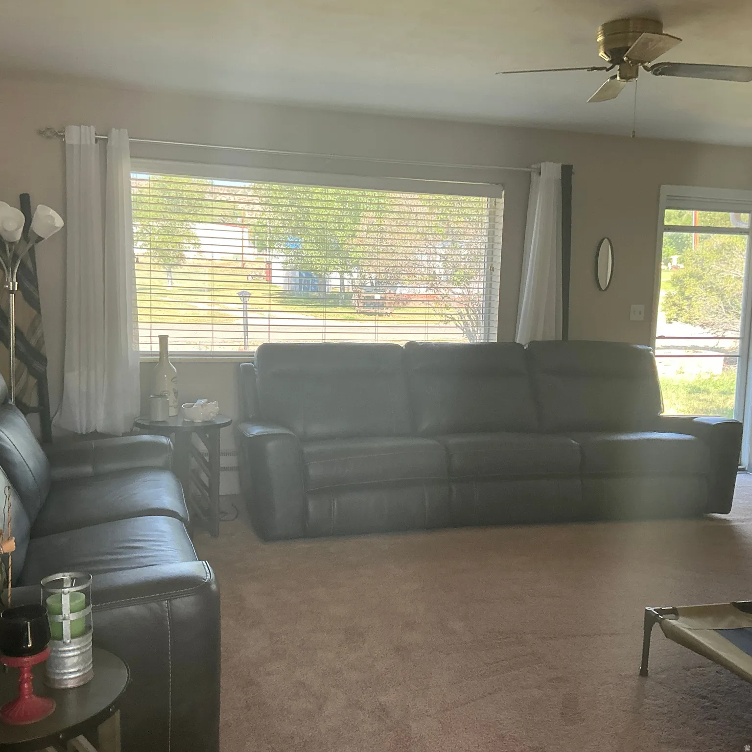 Living area featuring a large window with horizontal blinds, neutral-toned carpeting, and a ceiling fan with integrated lighting