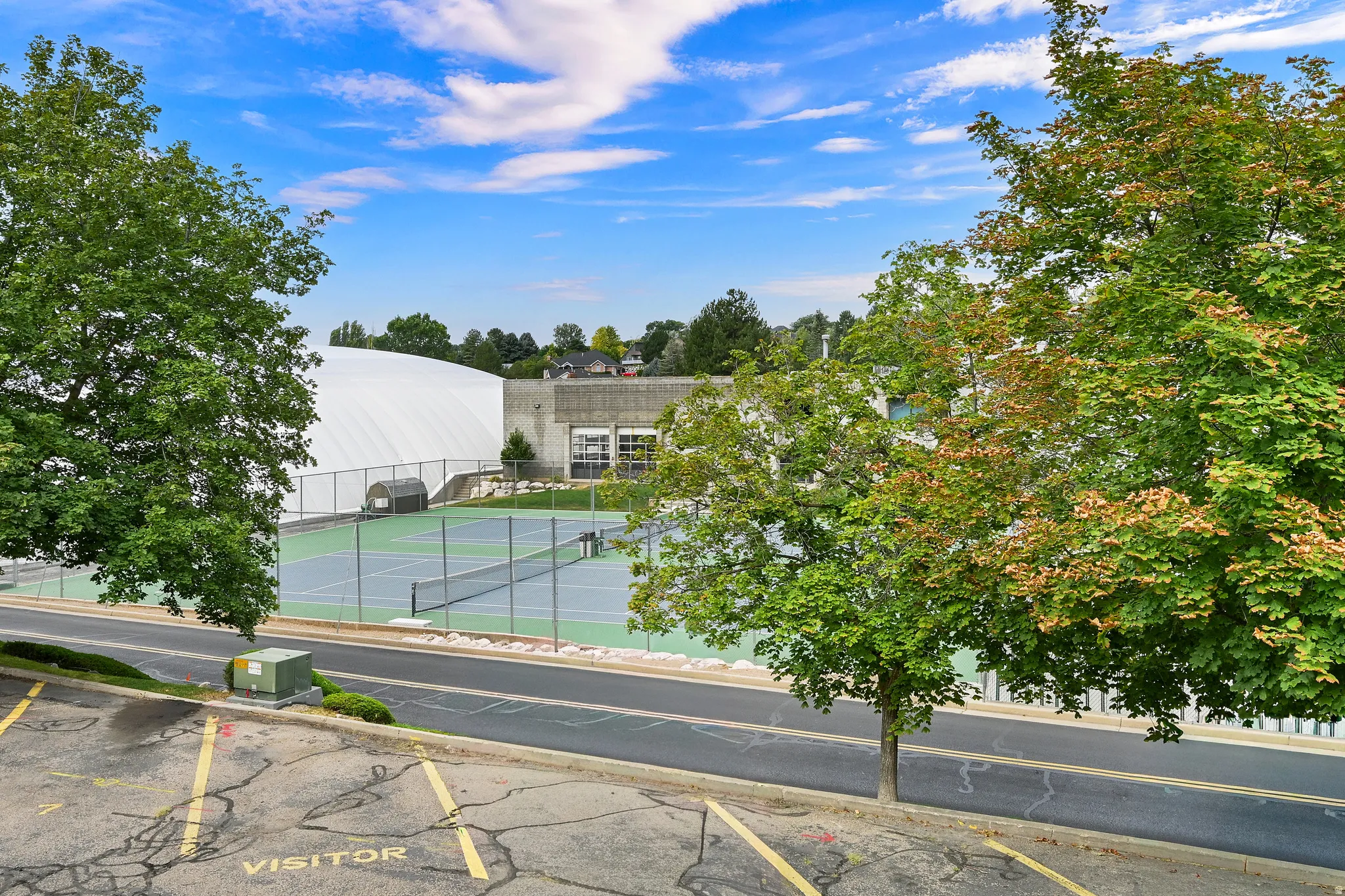 Outdoor tennis courts with chain-link fencing, surrounded by mature trees and a paved access road