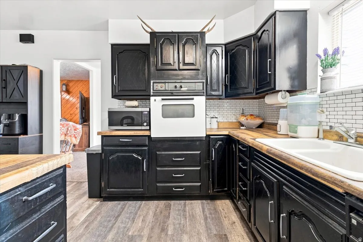 Kitchen featuring black cabinetry, wood-finish countertops, white subway tile backsplash, a built-in wall oven, and wood-finish flooring