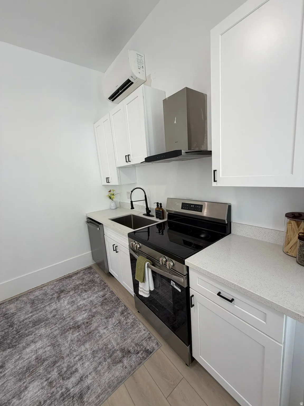 Contemporary kitchen featuring white shaker cabinetry with black hardware, light-toned countertops, a stainless steel range with overhead ventilation, and wood-finish flooring