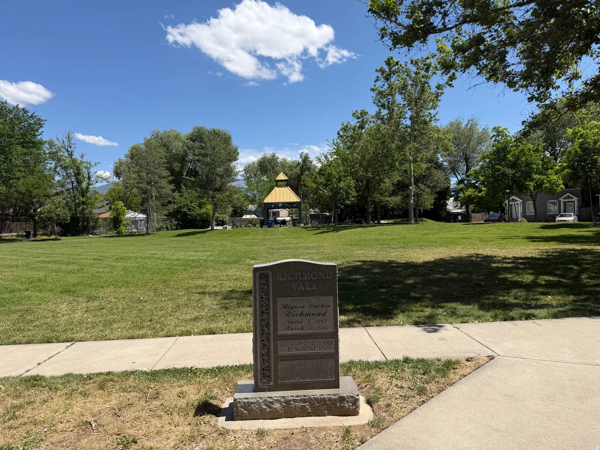 Expansive green space featuring a commemorative stone marker, concrete pathways, and mature trees