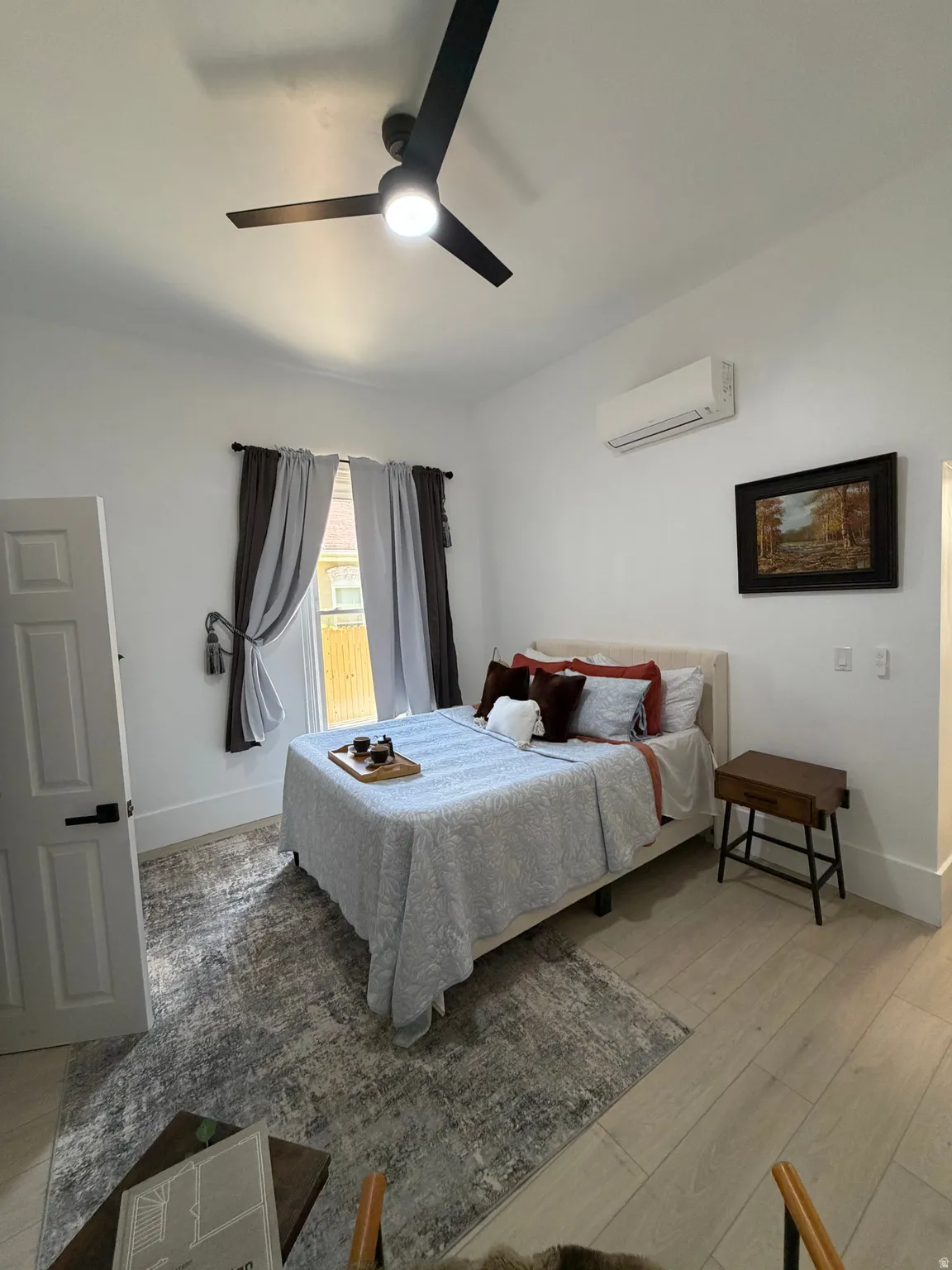 Bright room featuring light wood-finish flooring, white walls, a contemporary black ceiling fan with integrated lighting, and a wall-mounted mini-split HVAC unit
