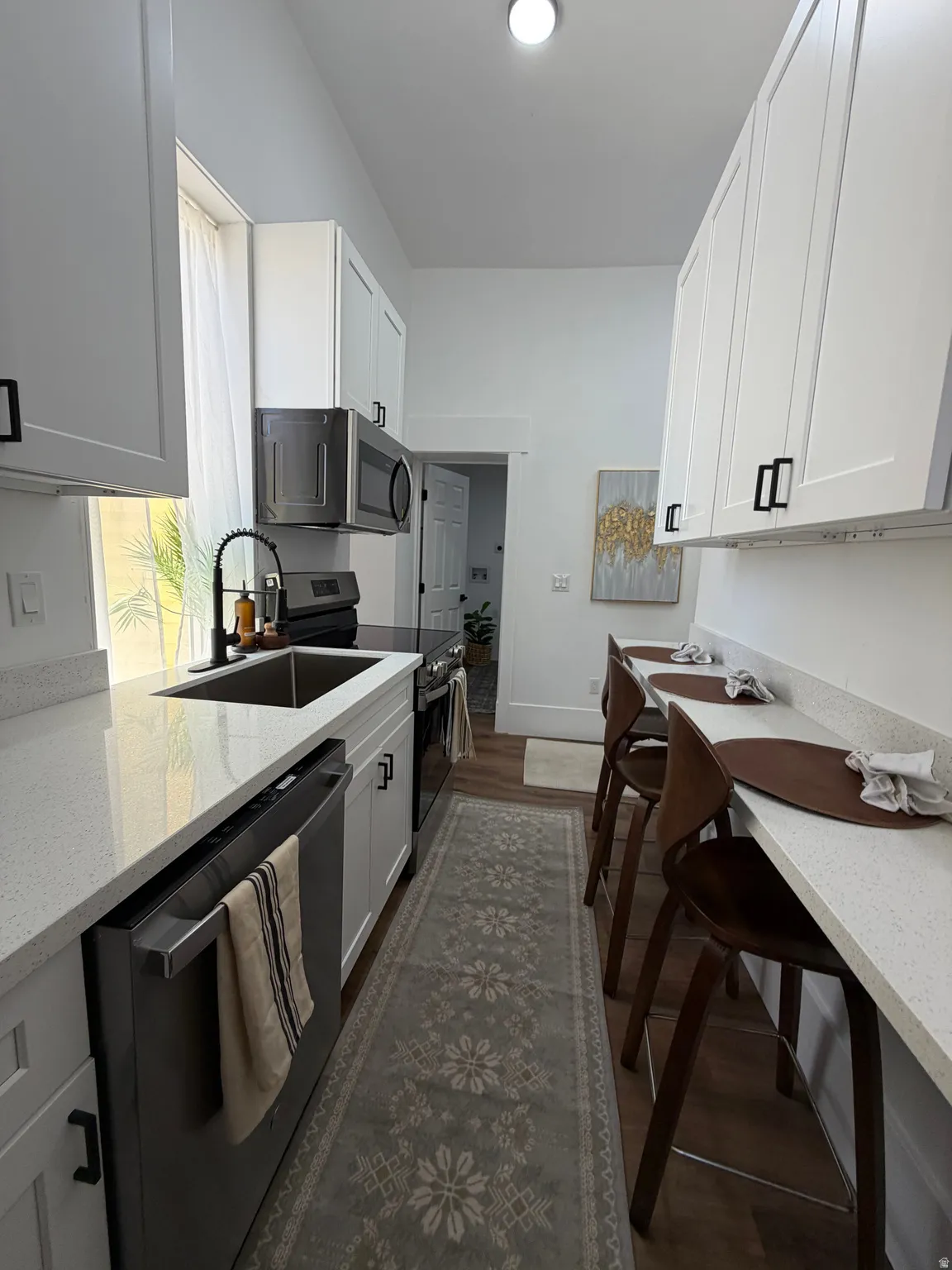 Galley kitchen featuring white shaker cabinetry with matte black hardware, stainless steel appliances, a bar-height dining counter, and wood-finish flooring