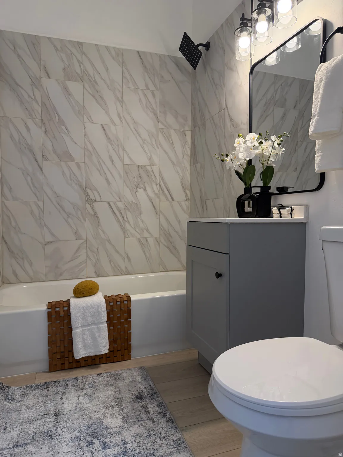 Full bathroom featuring a tub with a rain shower head, large-format marble-look wall tiling, a contemporary vanity with a white countertop, and wood-finish flooring